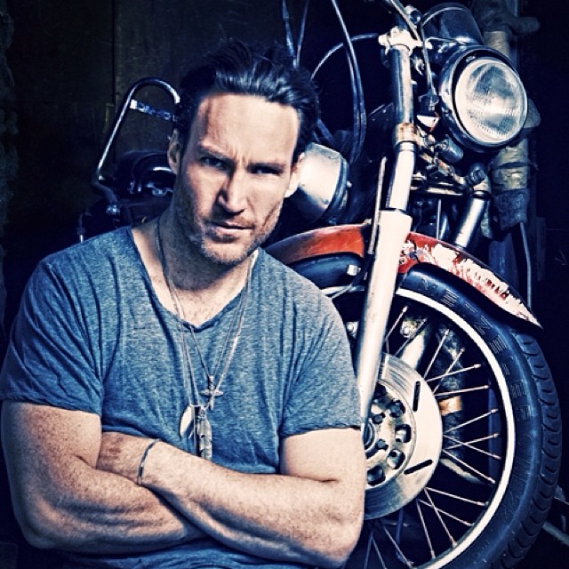 Callan Mulvey in a blue t-shirt folding his arms in front of his bike.